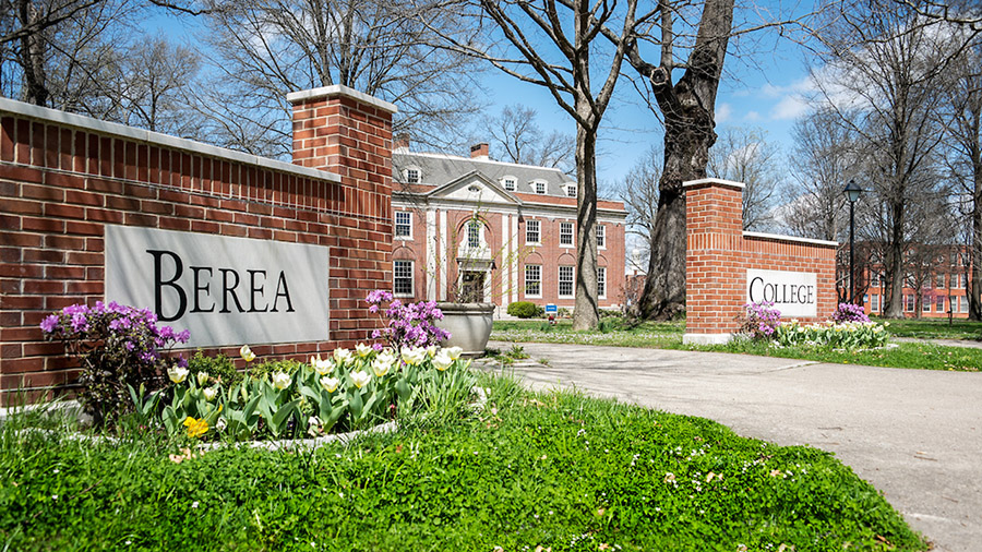Berea College entrance sign with brick pillars, flowering gardens, and historic red-brick buildings in background.