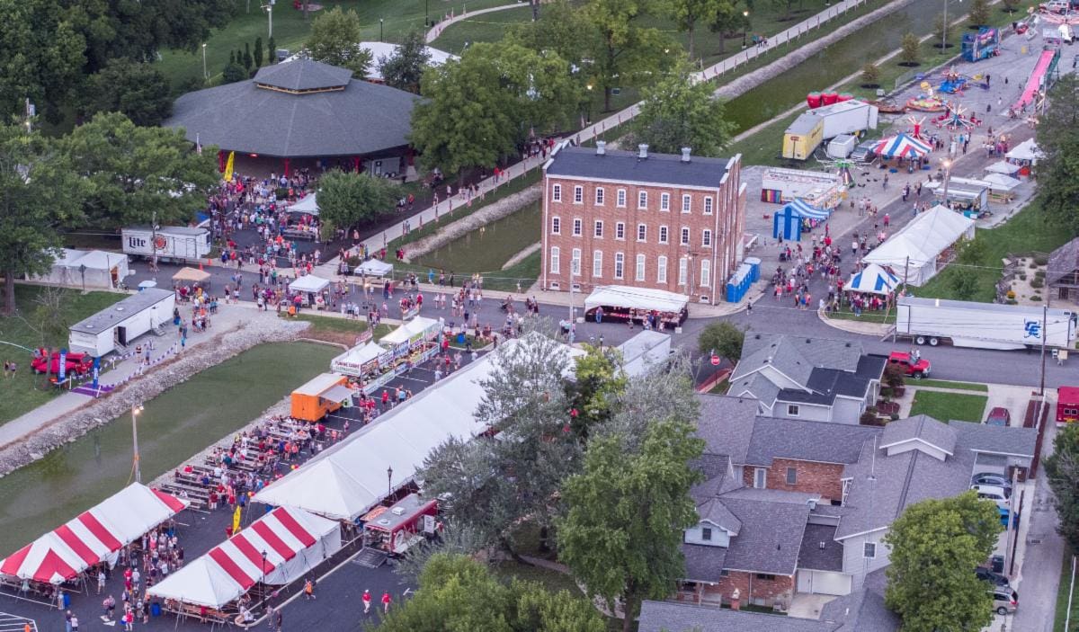 Aerial view of a festive street fair or carnival with red and white tents, crowds, and historic brick buildings.