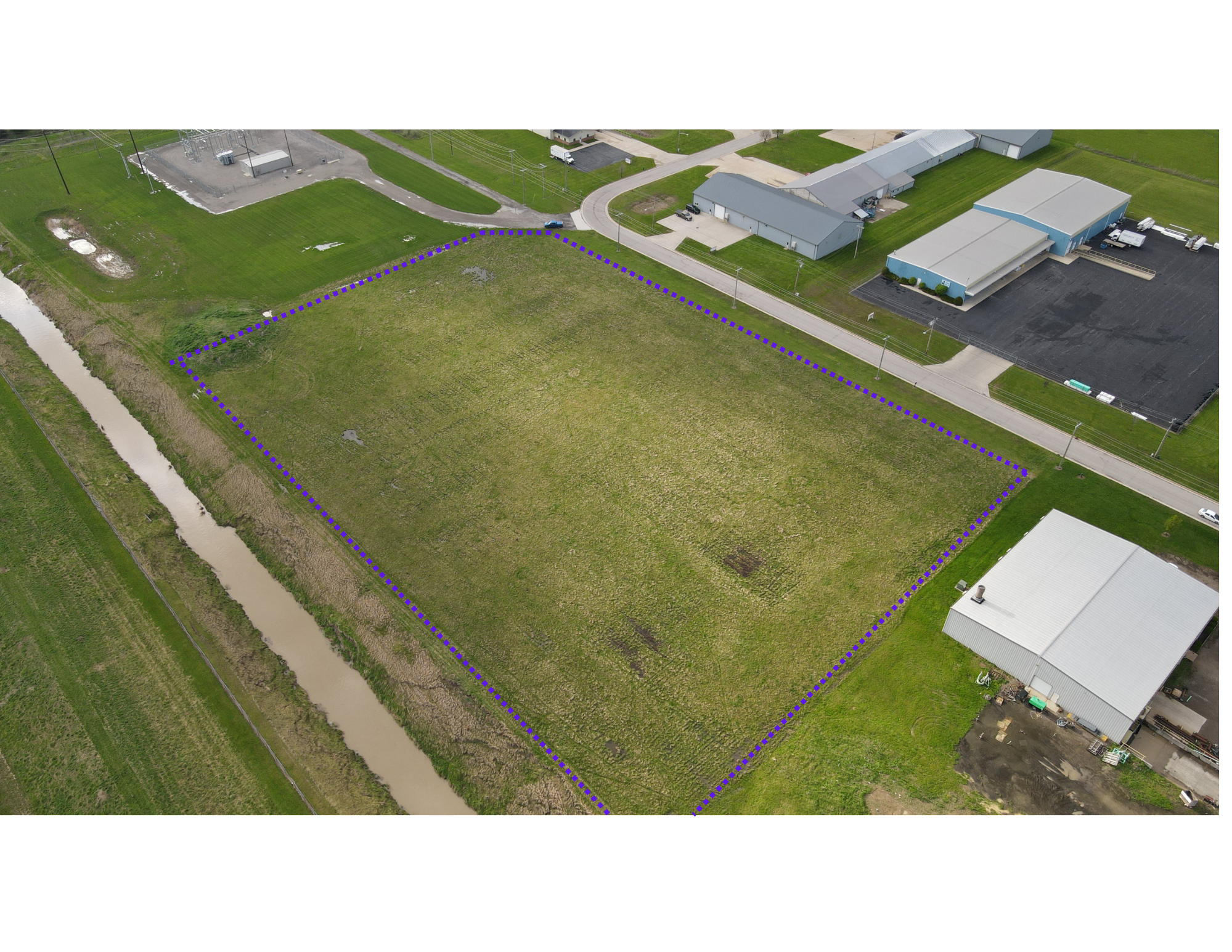 Aerial view of a vacant rectangular lot with grass and border markings, adjacent to industrial buildings and a canal.