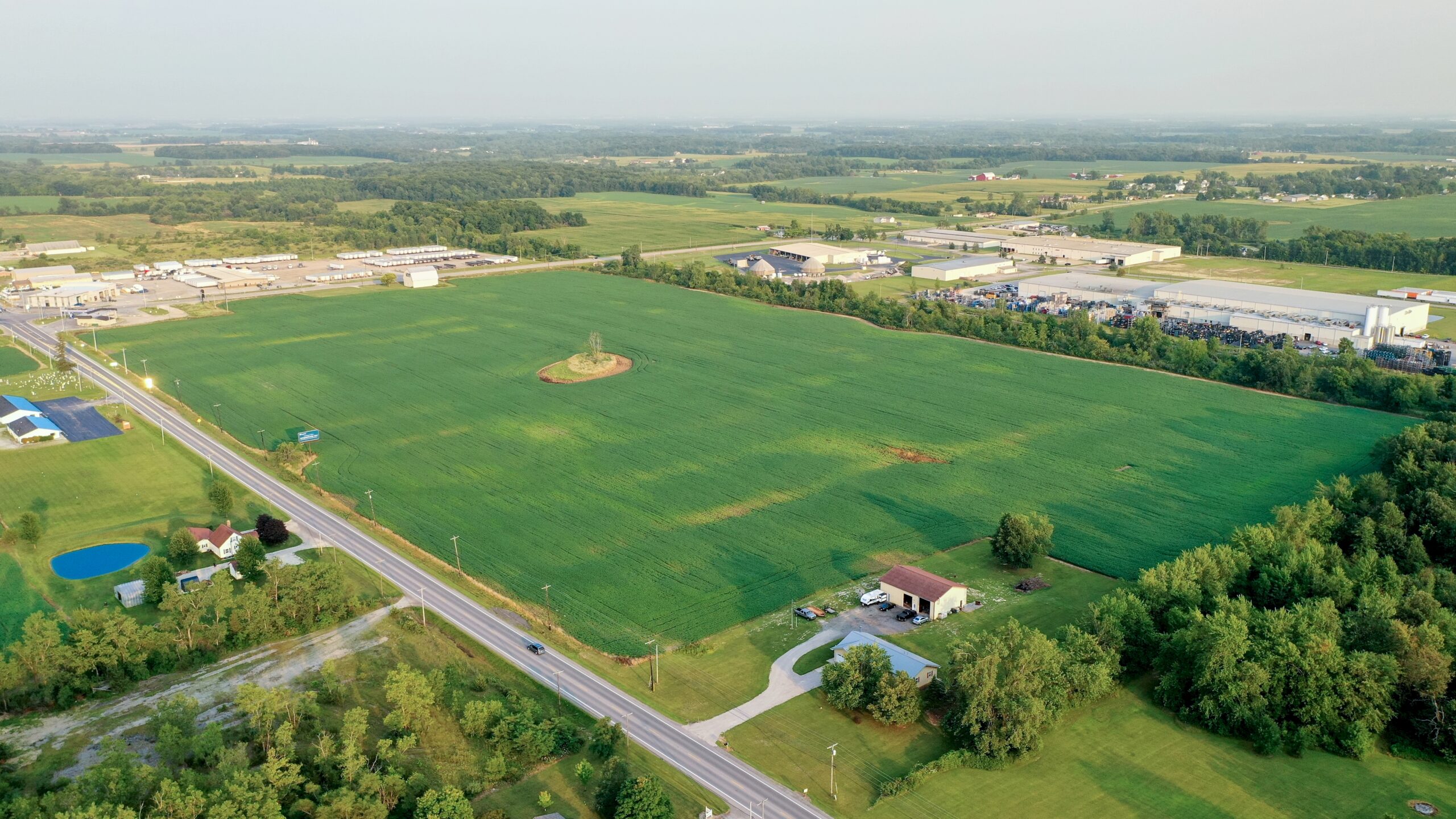 Aerial view of a sports complex with baseball fields, a straight road, and surrounding agricultural land and forests.