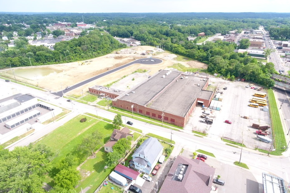 Aerial view of an industrial facility with large warehouse buildings, parking areas, and surrounding green landscape and residential neighborhoods.