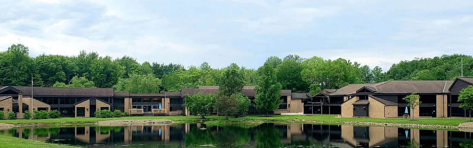 Modern brick buildings reflected in a calm waterfront pond surrounded by green trees.