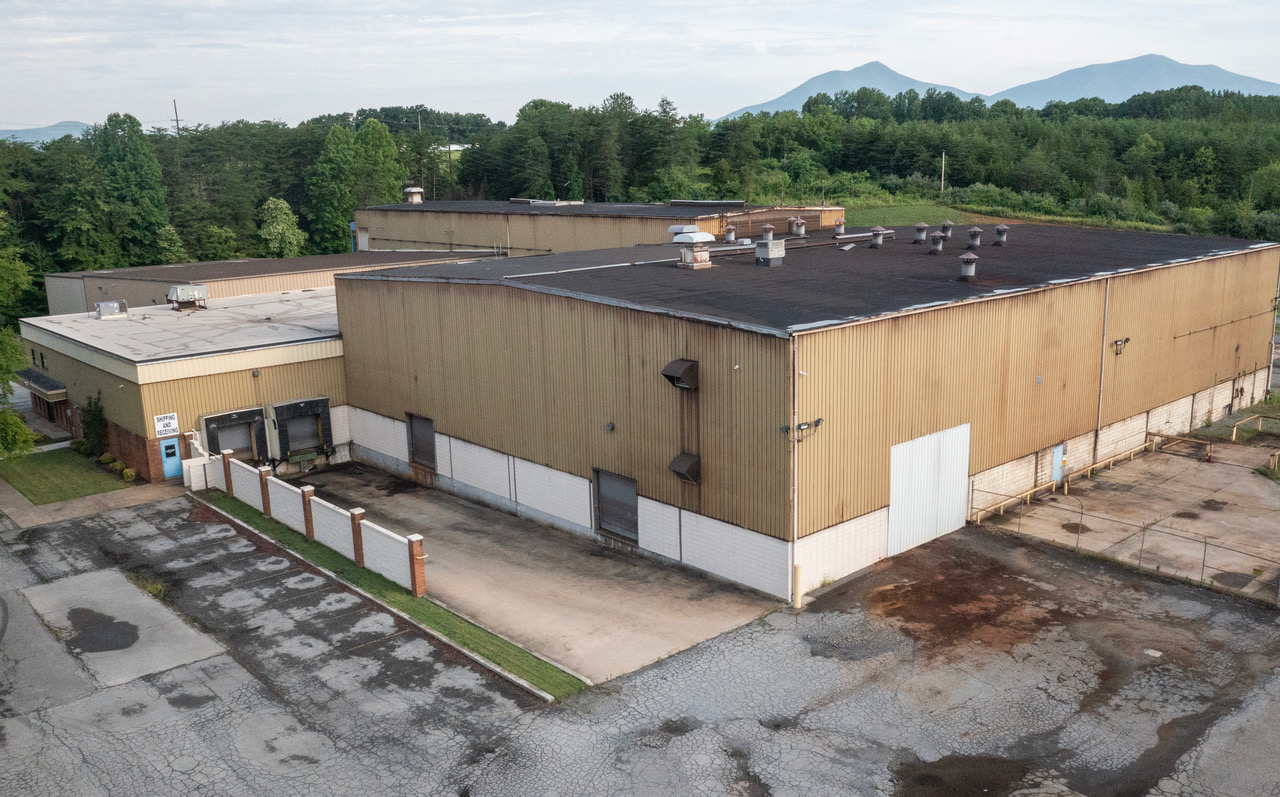 Aerial view of a large industrial warehouse building with tan metal siding and dark roof in a forested area with mountains in the distance.