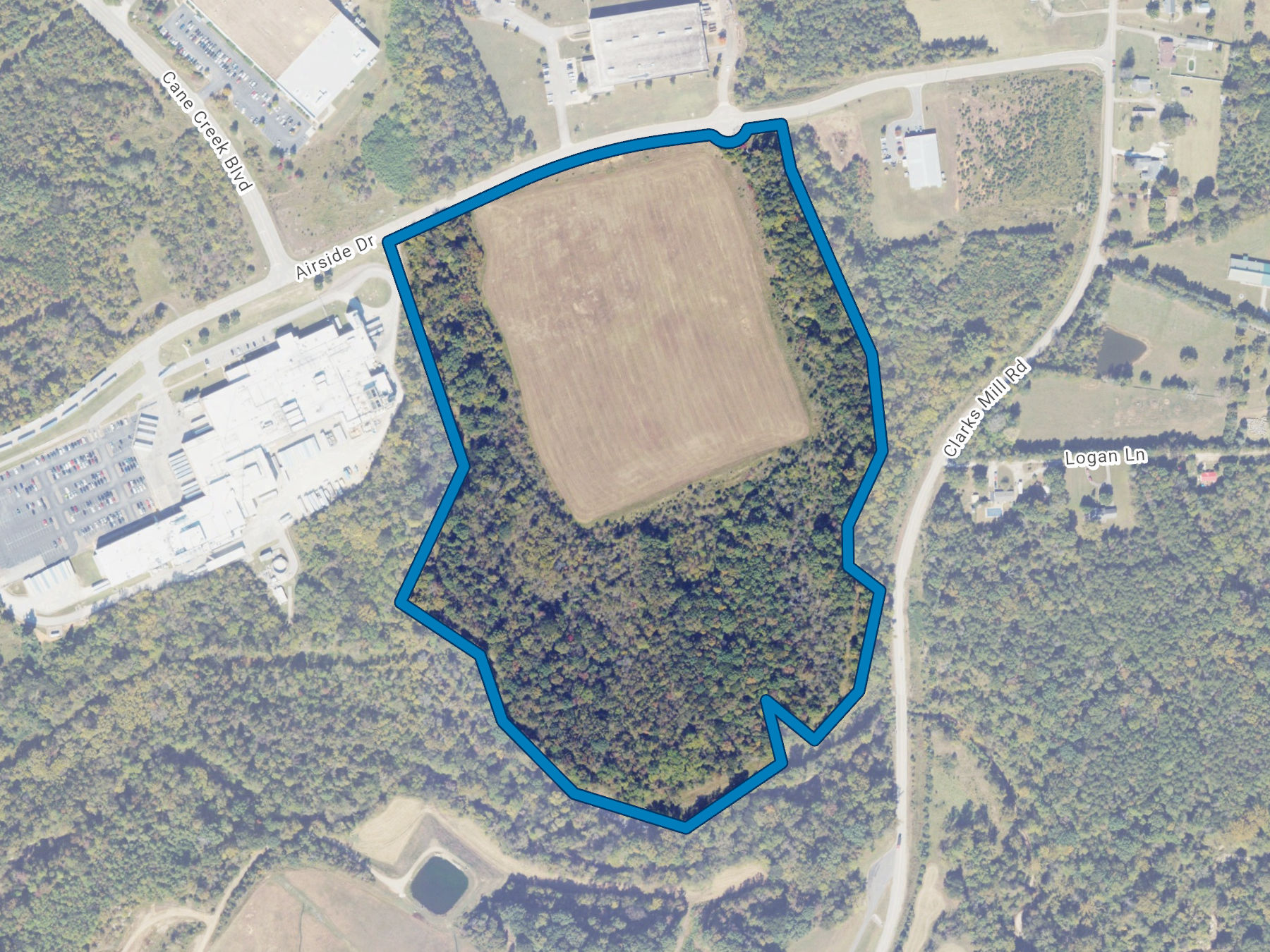 Aerial map view of a property outlined in blue, showing cleared land surrounded by forest and adjacent industrial buildings.