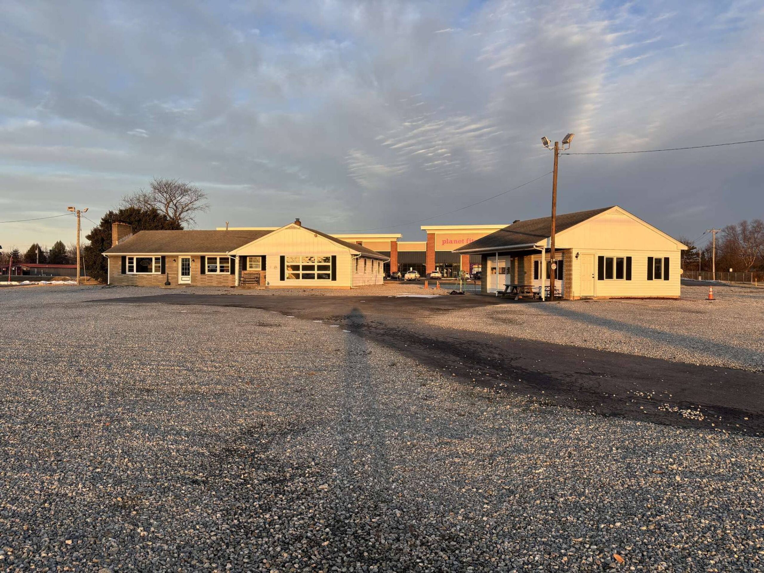 Yellow commercial buildings on a gravel lot with bare trees and utility poles under a cloudy sky.