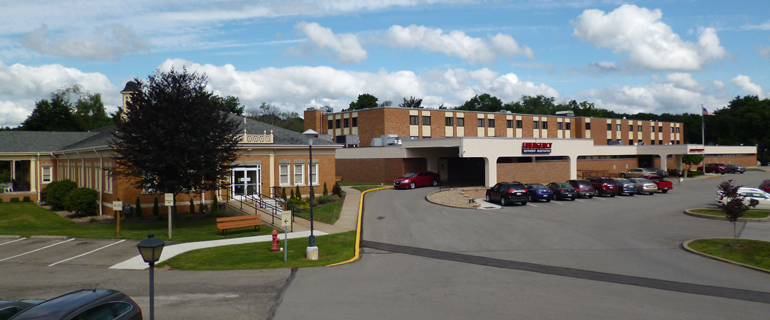 Exterior view of a brick commercial or medical building with parking lot and landscaped entrance area under blue sky.