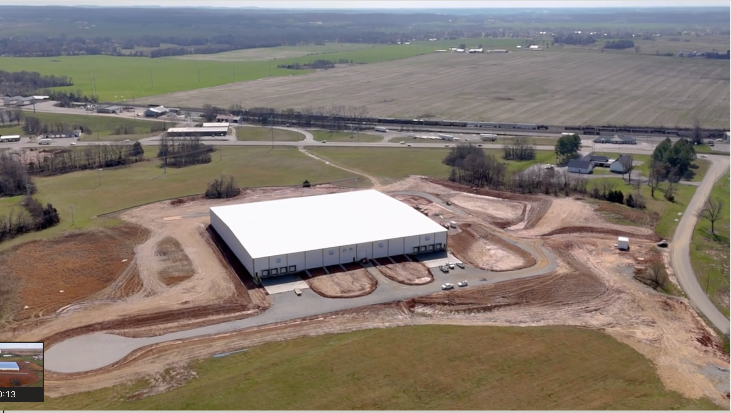 Aerial view of a large industrial warehouse under construction with white roof, surrounded by earthwork and rural farmland.