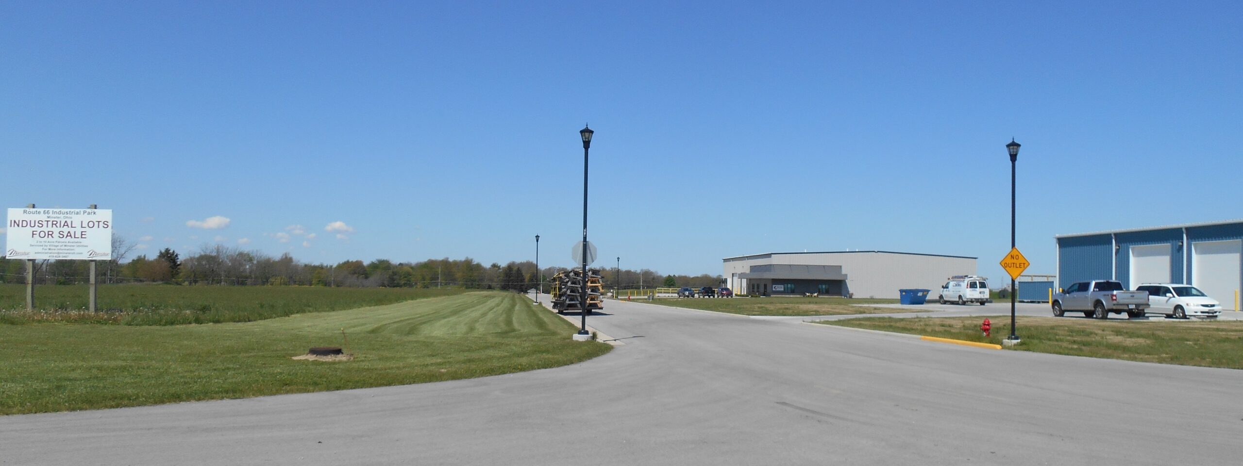 Industrial park entrance with paved road, green fields, commercial buildings, and clear blue sky.