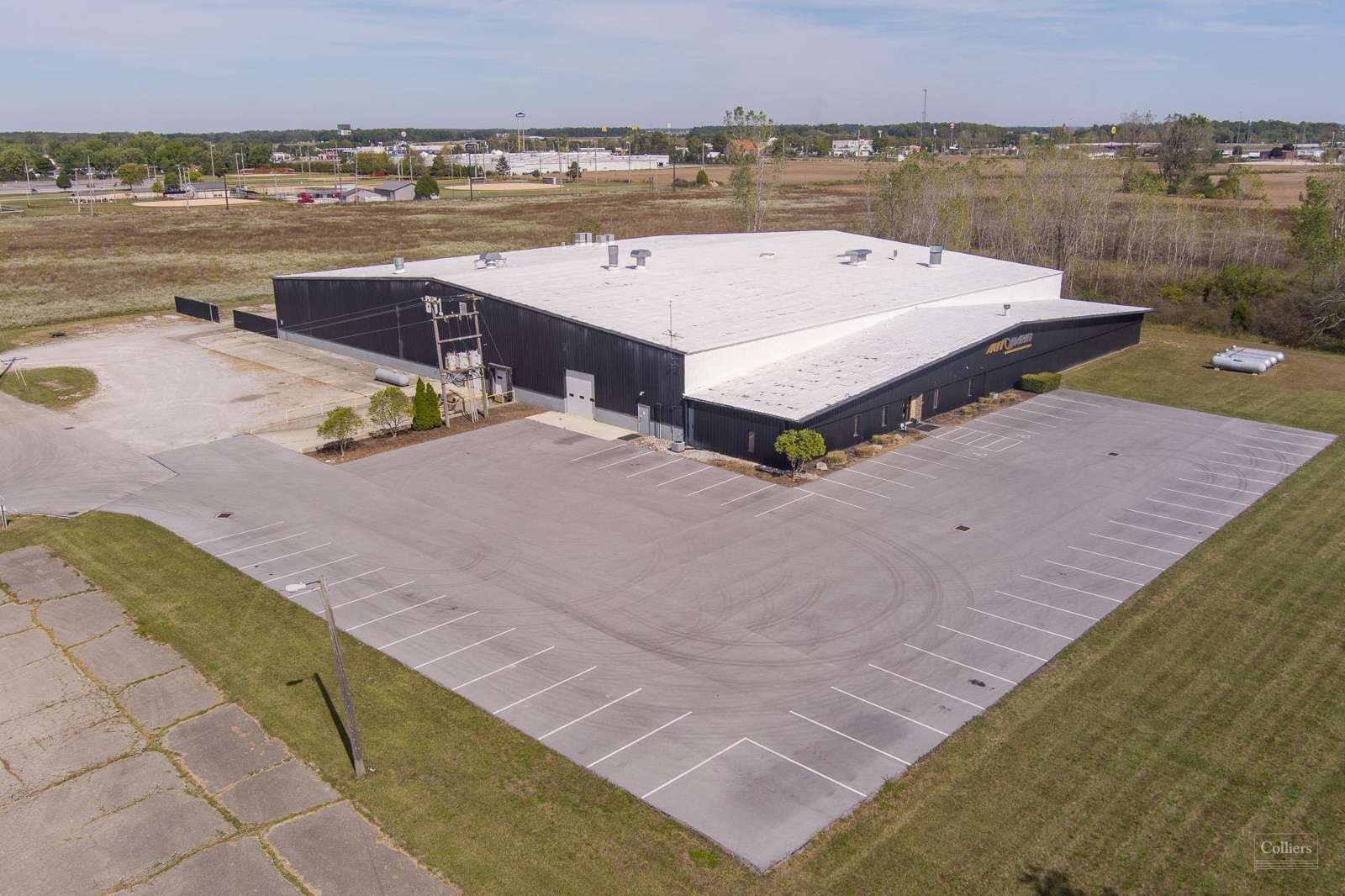 Aerial view of a modern industrial building with white roof and black walls on a large paved lot surrounded by grassland.