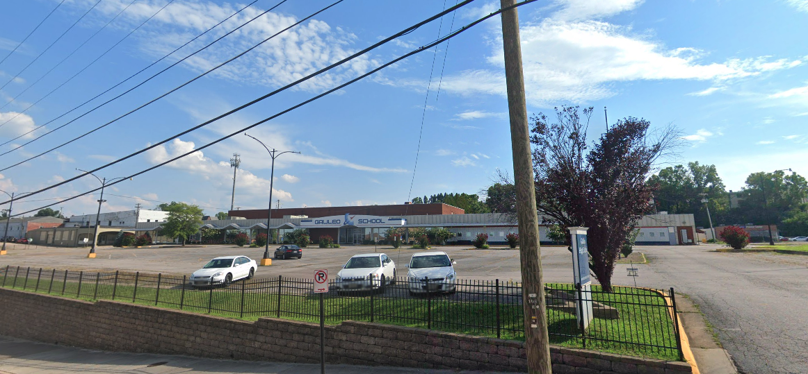 Car dealership lot with vehicles displayed behind black fencing along a commercial street.