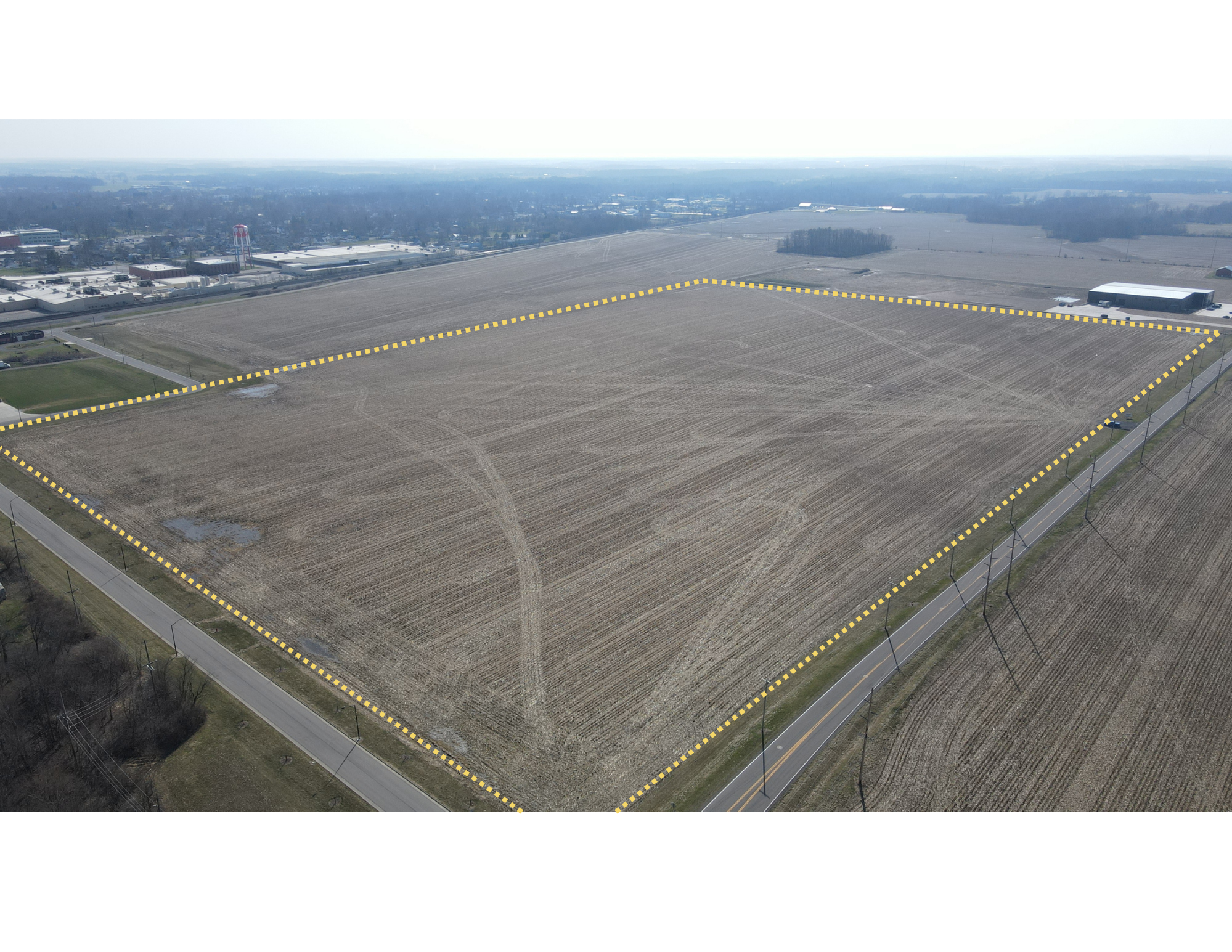 Aerial view of a large rectangular agricultural field outlined in yellow, bordered by roads and surrounded by farmland and industrial areas.