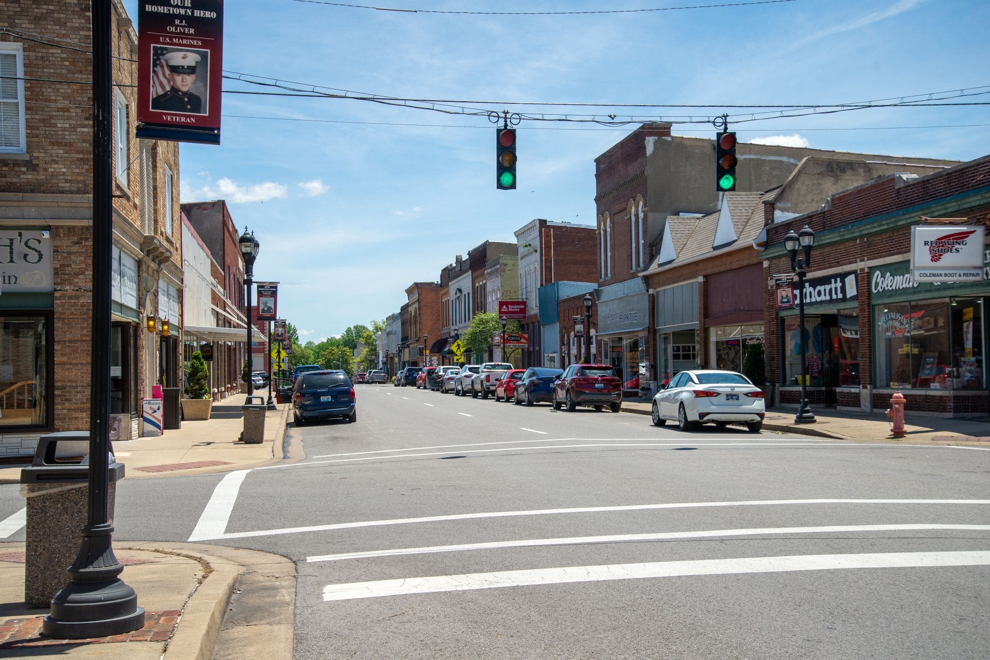 A sunny downtown street with brick buildings, parked cars, traffic lights, and storefronts lining both sides.