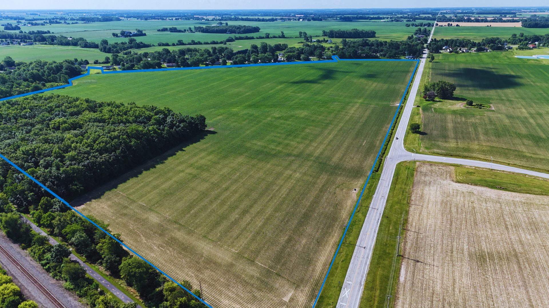 Aerial view of rural farmland with green fields, forests, roads, and scattered buildings in a flat agricultural landscape.
