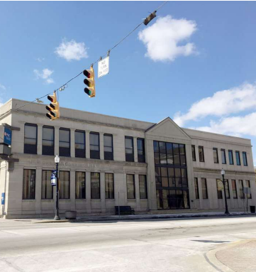 A modern gray institutional building with large windows and glass entrance, situated on an empty plaza with traffic lights overhead.