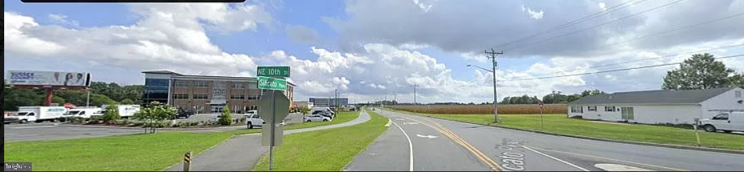 A wide road with commercial buildings and green fields under a partly cloudy sky.