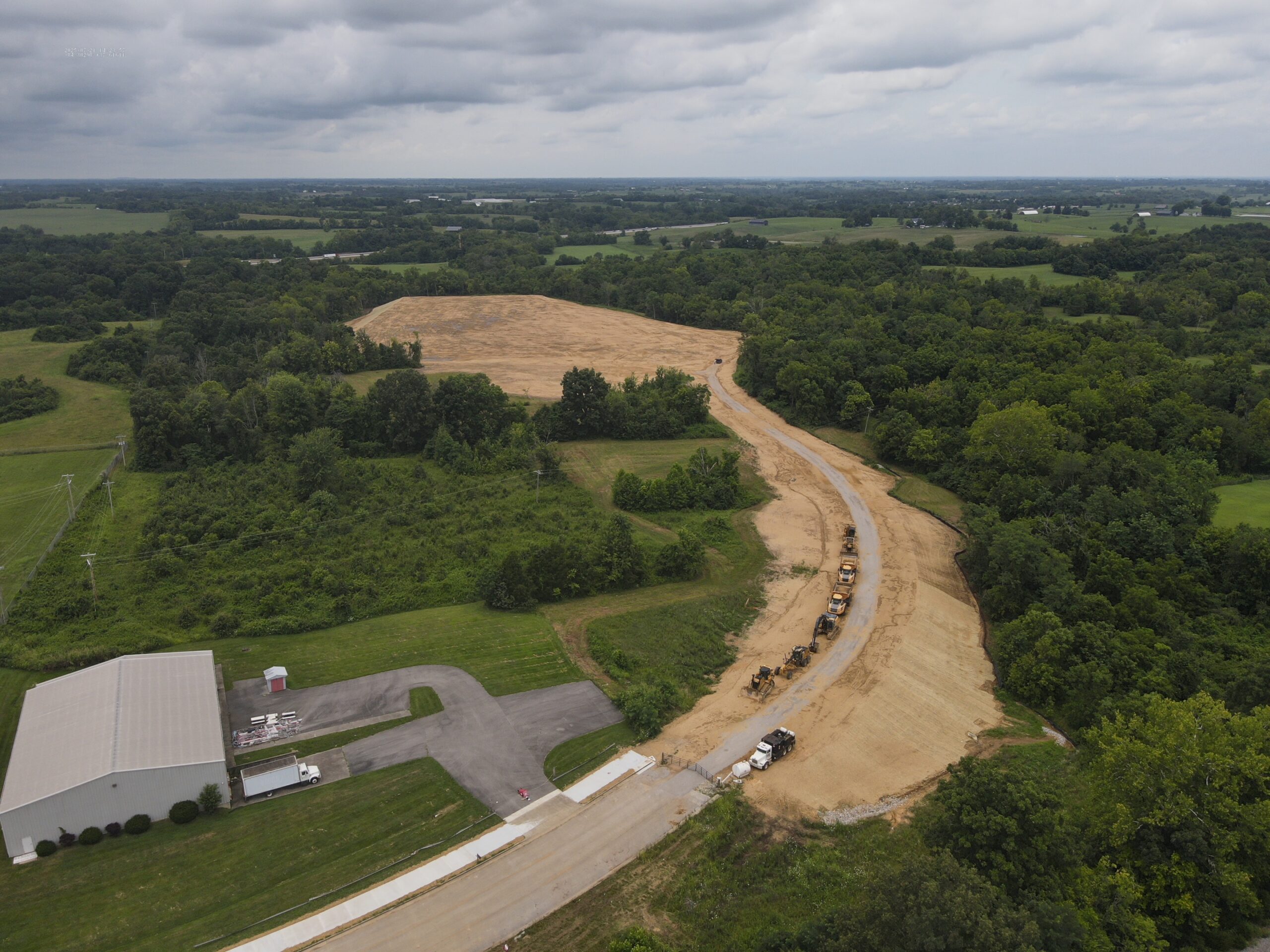 Aerial view of a curved dirt road construction project cutting through forested farmland with a metal warehouse building nearby.