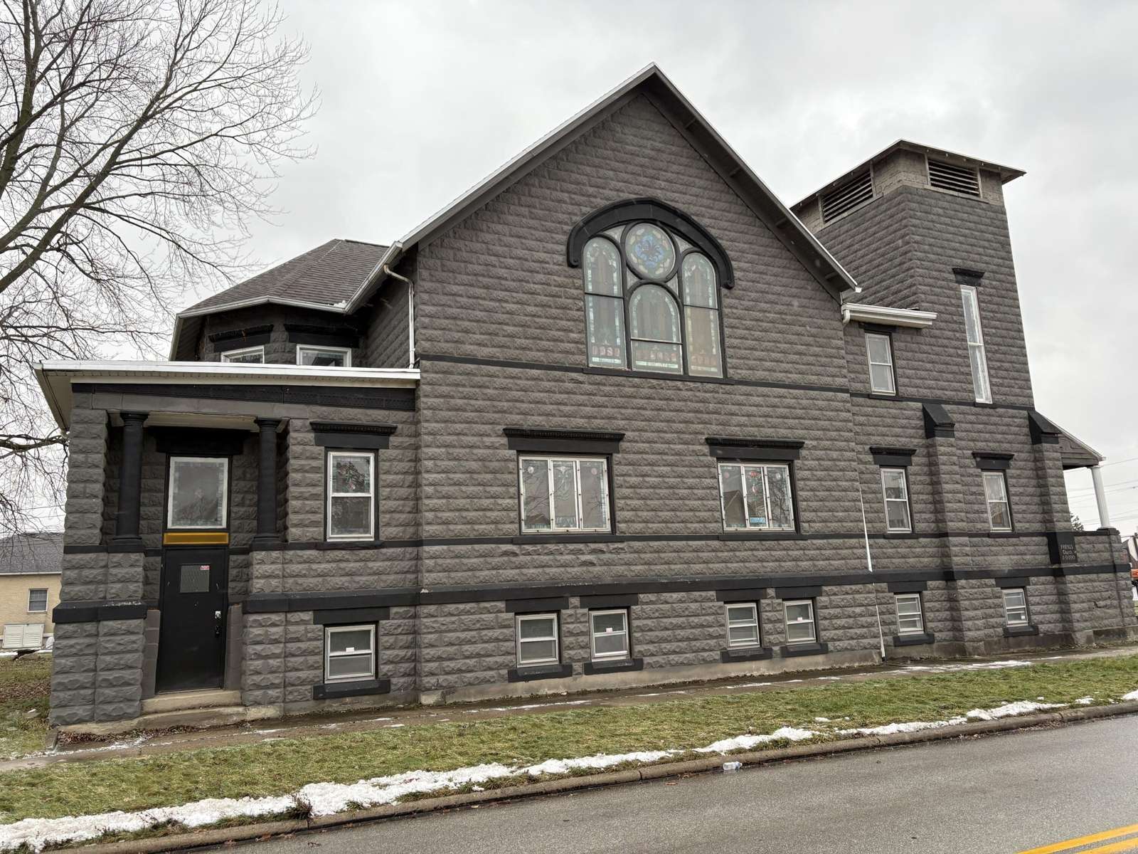Gray Victorian-style house with peaked roof, arched window, and tower, situated on a sloped lawn.