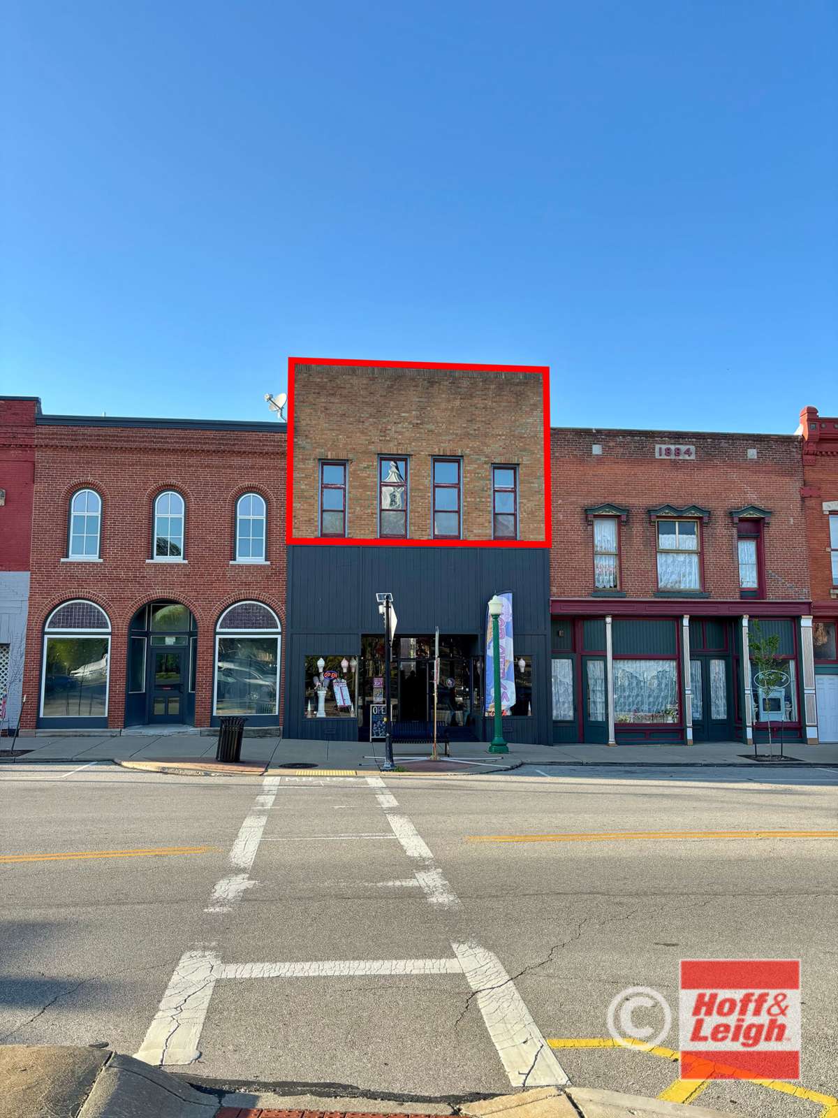 Historic brick storefronts on a sunny street with a red-framed building in the center and white crosswalk markings.