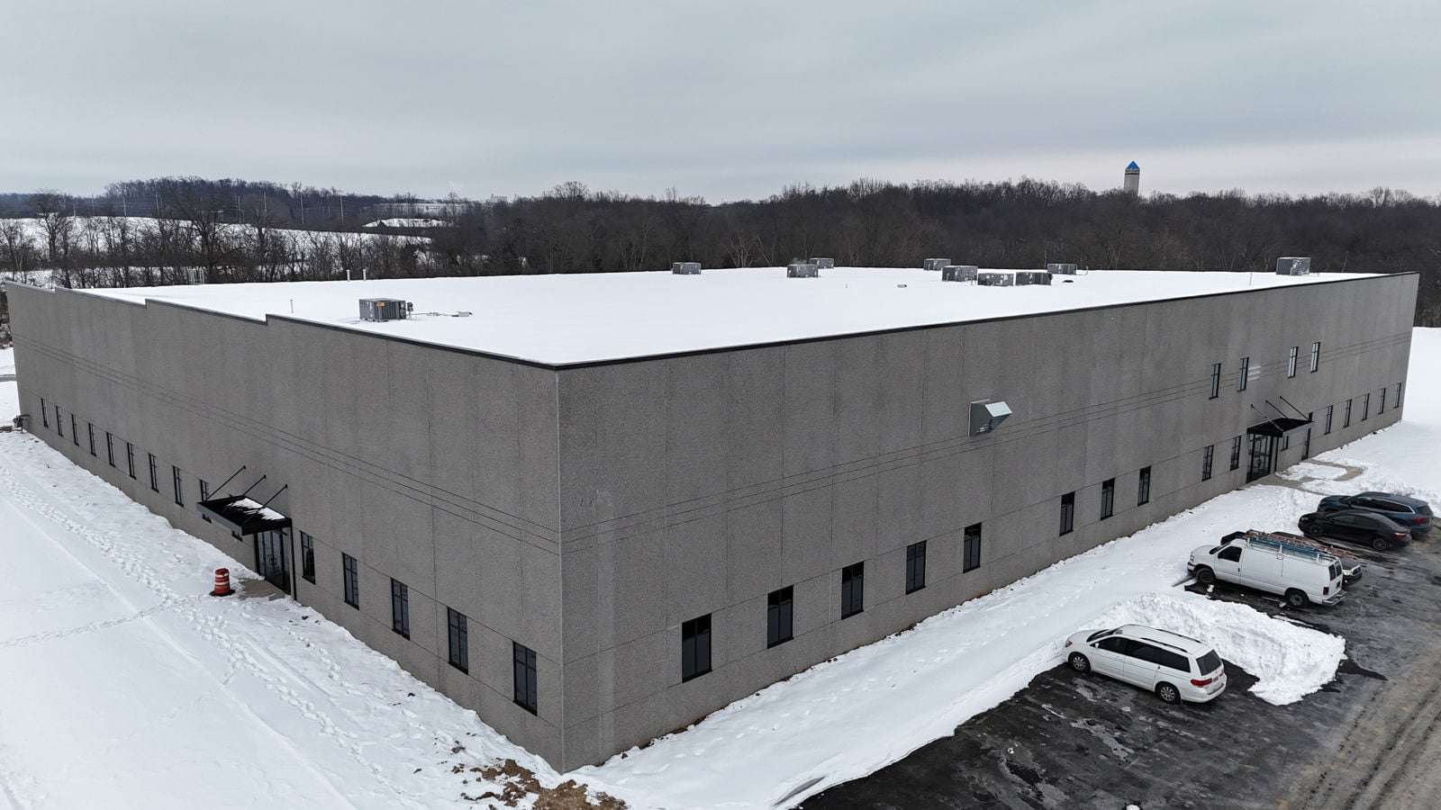 Modern industrial building with white roof and gray concrete walls in snowy setting with parked vehicles.