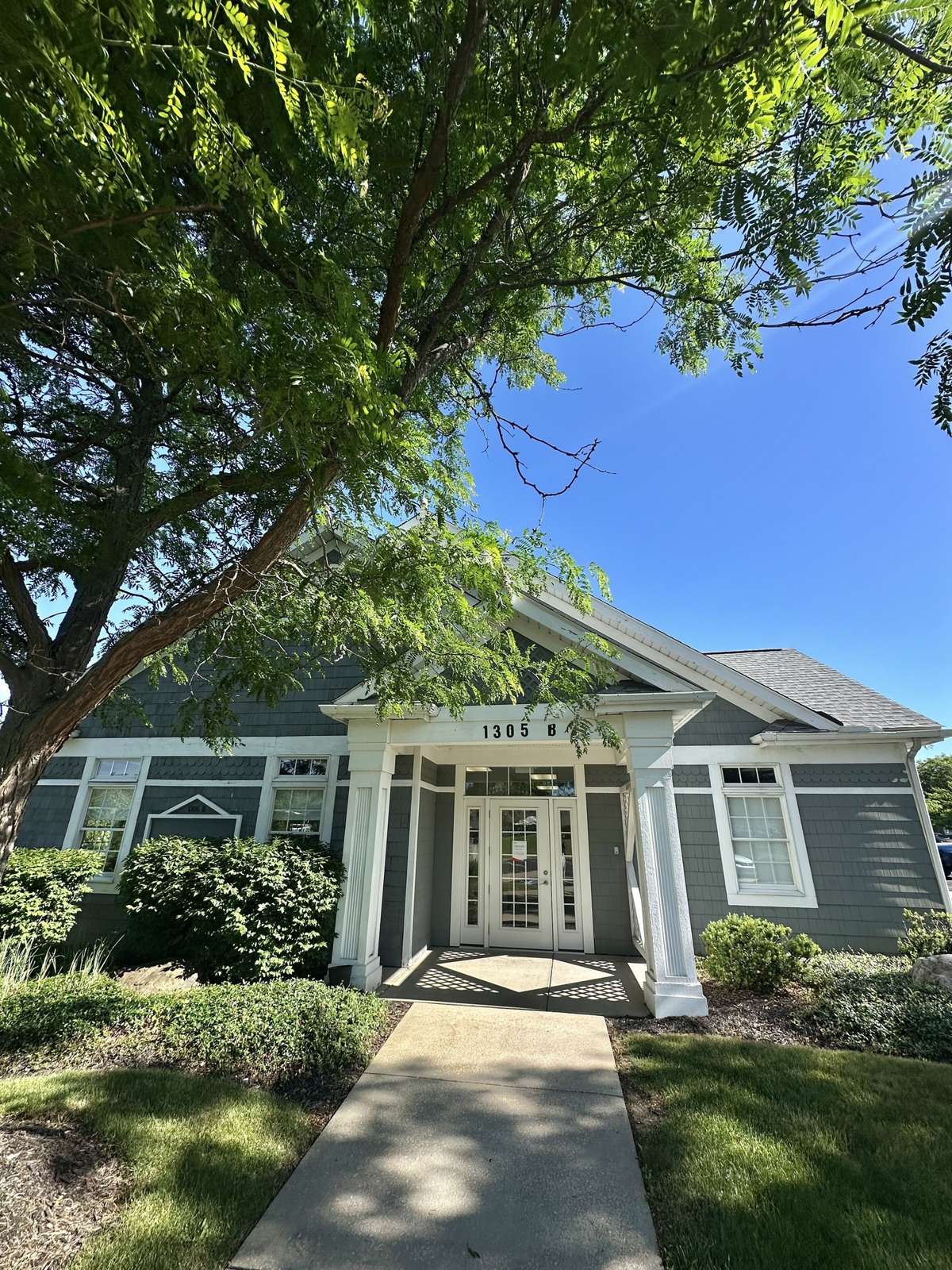 Front exterior of a gray and white single-story house with columns and large shade tree.