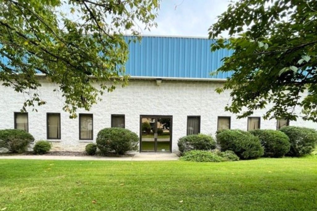 Modern white brick building with blue metal roof and landscaped grounds, featuring large windows and manicured lawn.